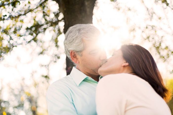 elderly couple kissing outdoors, sun shining through trees in background