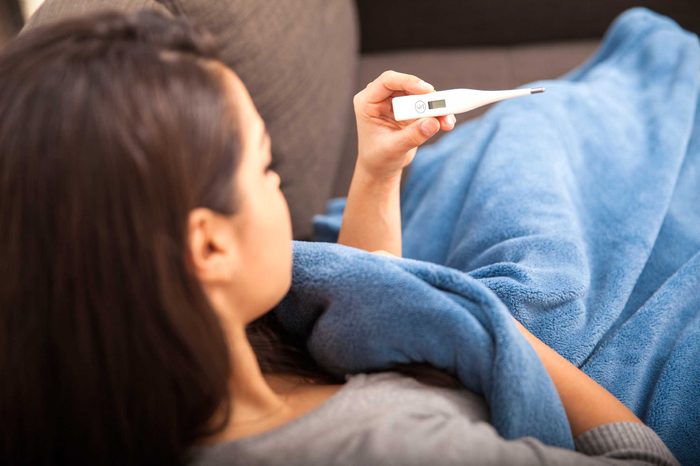 Woman sitting on a couch covered in a blanket holding a thermometer in her left hand.