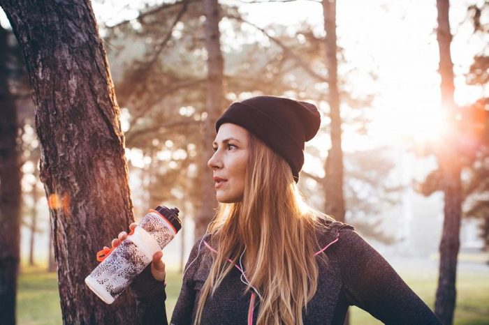 Woman runner wearing a beanie cap and drinking out of a water bottle while standing in a park.