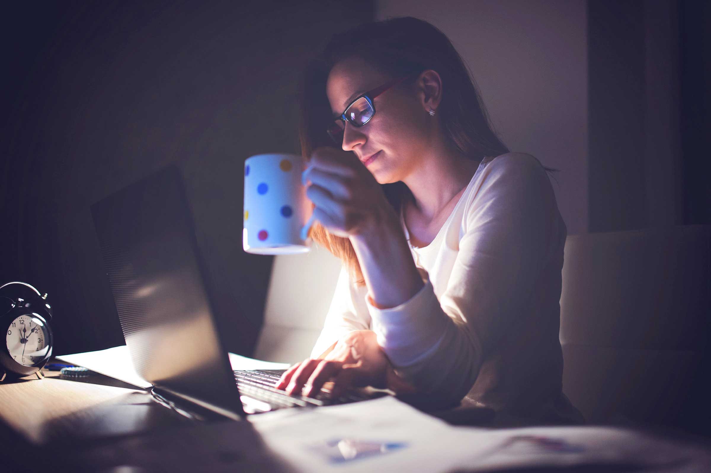 woman holding a cup of coffee and working at a computer in a dark room