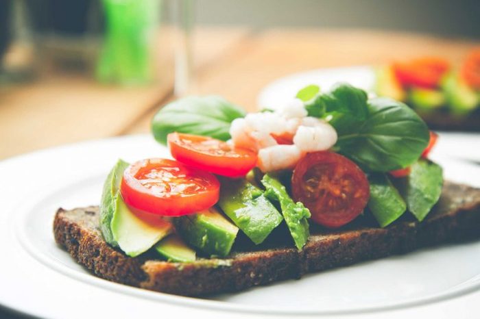 Vegetables like tomatoes and cucumbers piled on a piece of bread that's sitting on a plate.