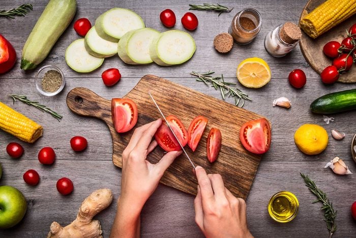 person cutting tomato on chopping board, surrounded by fresh vegetables