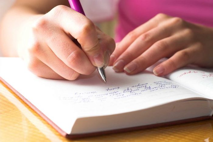 Woman in a pink top writing in a journal with a pen.