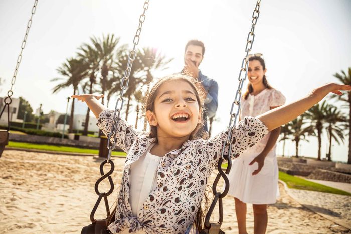 Little girl sitting on a swing in a park with her parents in the background.