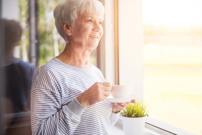 Smiling older woman enjoying a cup of tea while peering out a window.