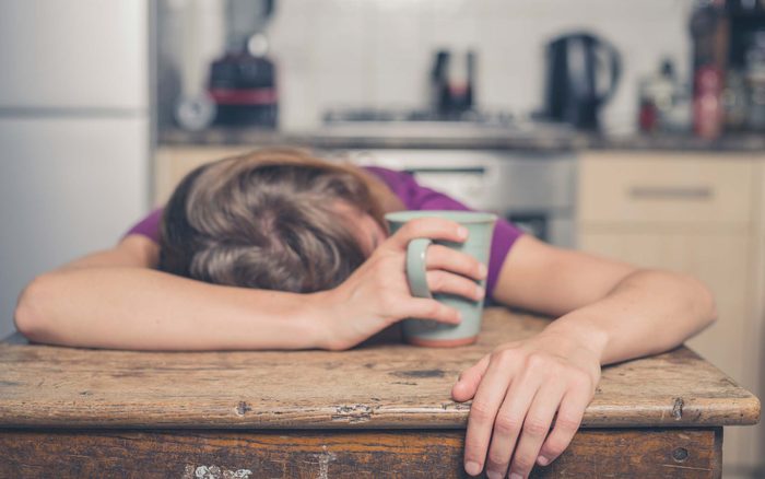woman collapsed on a wooden kitchen table holding a mug