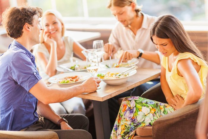 A family sitting around a table eating a meal together, while one woman is clutching her stomach as if in pain.