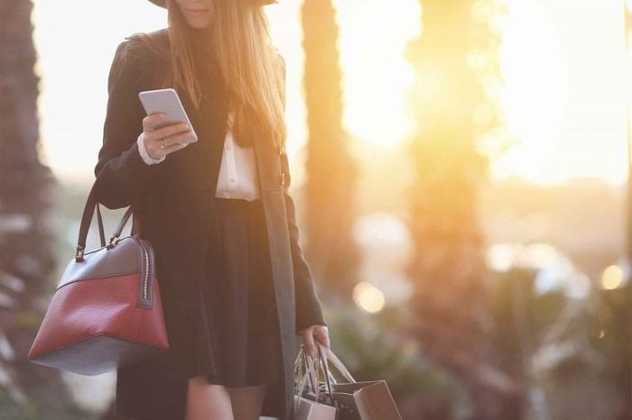Woman wearing trendy clothing carrying shopping bags and looking at her phone as the sun sets behind her.