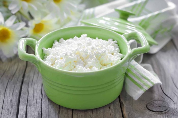 Green bowl of cottage cheese sitting on a wooden tabletop near a bunch of white flowers and a white and green cloth napkin.