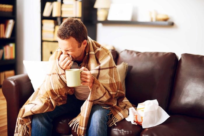 man wrapped in a blanket on a couch, sneezing