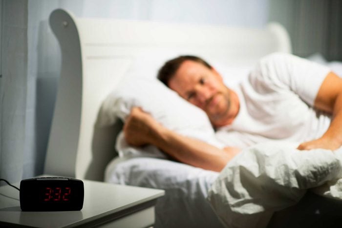 awake man lying on bed looking at a bedside table clock