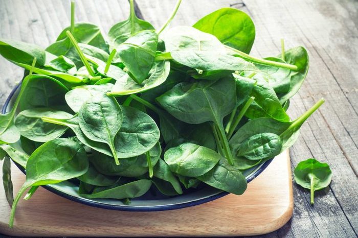 Bowl of leafy green spinach in a blue bowl resting on a wooden cutting board and wooden tabletop.