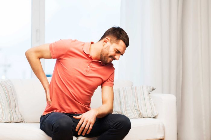 Man sitting on the couch holding his back as if having a backache.