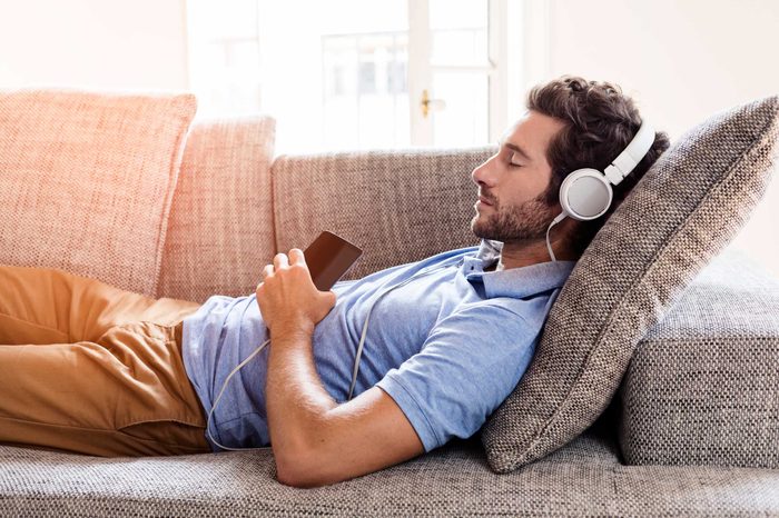 Man closing his eyes, reclining on the couch while wearing headphones and listening to music.