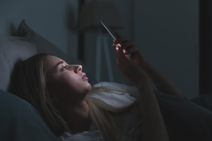 Young sleepy woman lying in bed using smartphone