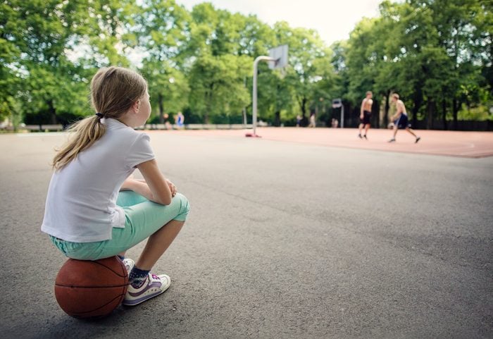 young girl sitting on basketball watching people play