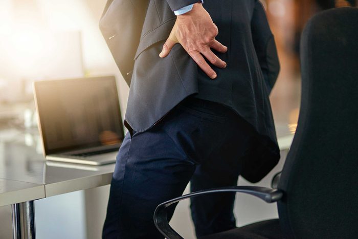Man in a blazer standing up from an office chair and grabbing his lower back