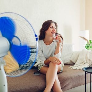 woman sitting on couch in front of fan and drinking a glass of water