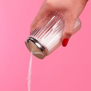 close up of hand pouring sugar out of container against a pink background