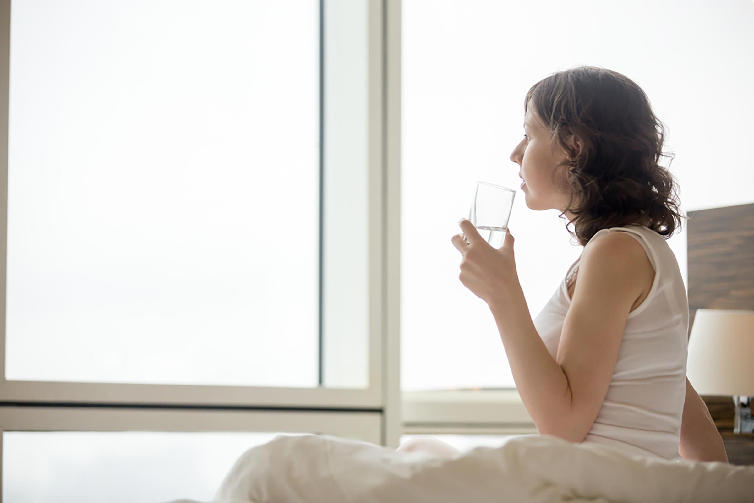 Woman sitting on edge of bed drinking glass of water