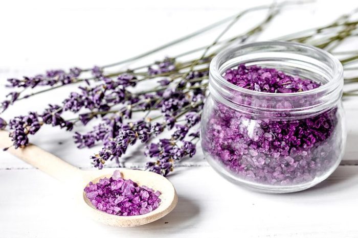 lavender stems on a white surface next to a wooden spoon and clear jar holding lavender petals