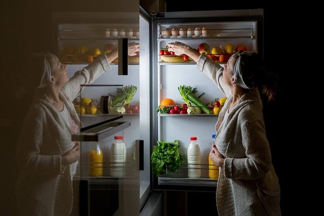 woman at open refrigerator snacking at night