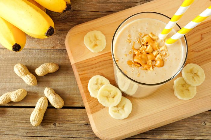 smoothie in a tall glass surrounded by banana slices, bananas and peanuts in the shell