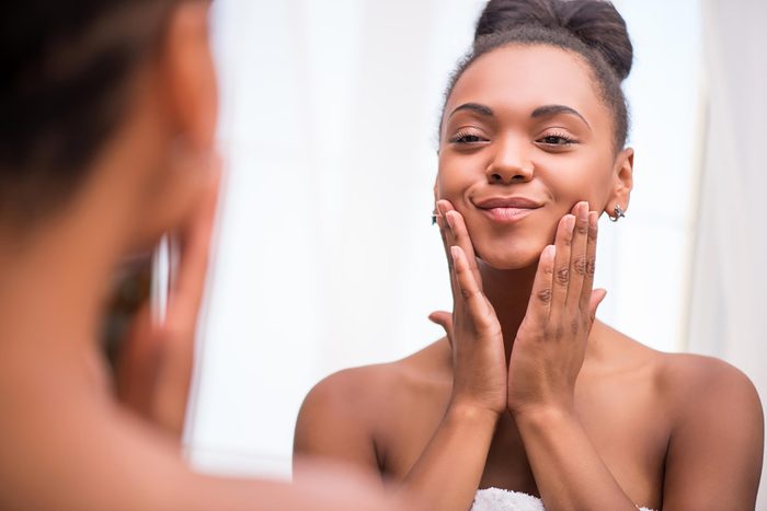 black woman smiling and examining her complexion in the mirror