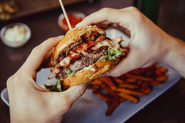 Hands holding a bacon cheeseburger over plate of fries