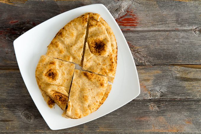 Overhead view of a crisp golden Naan flatbread baked in a traditional tandoor served on a modern square shaped white plate sliced into quarters on a rustic wooden table