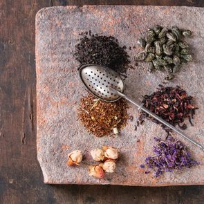 Variety of black, green, rooibos, herbal dry tea leaves and rose buds with vintage strainer on terracotta board over old dark wooden background. Top view with copy space