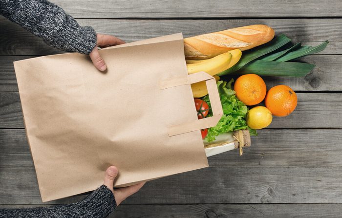 mãos segurando um saco de papel de mantimentos, incluindo legumes, frutas e pão