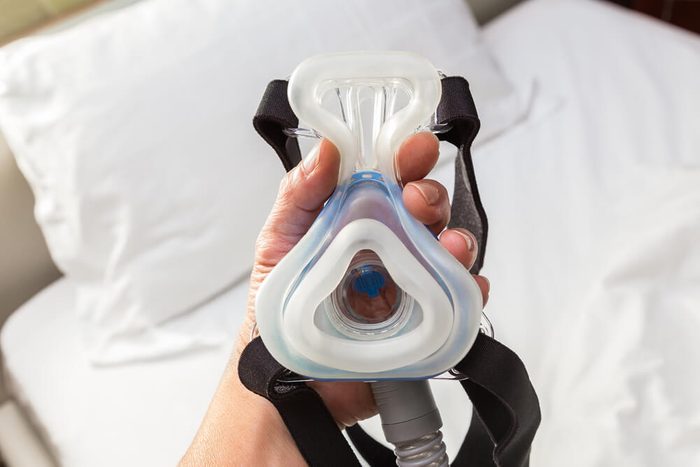 male hand holding CPAP mask, connecting with strap and hose, above his bed, wellness and respiratory