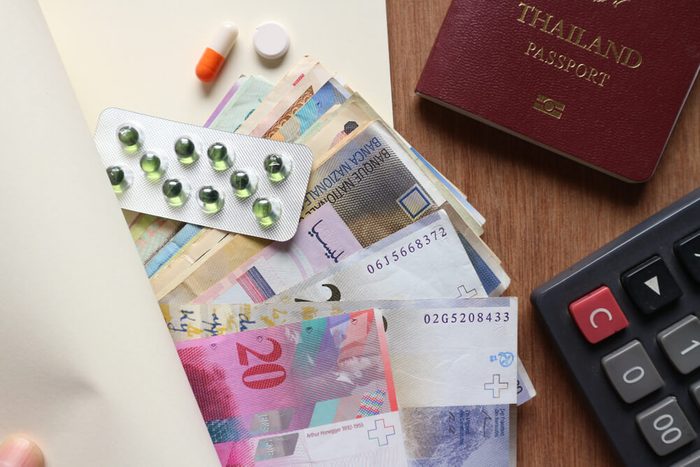 Various money with medicine panel, pill and capsule inside blank notebook with passport and calculator on wood table, finger on edge of book