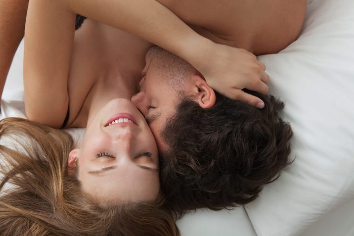 Overhead close up portrait of a young romantic couple hugging and kissing, laying down on a white bed, having sex and loving each other. Love and relationships lifestyle, interior bedroom.
