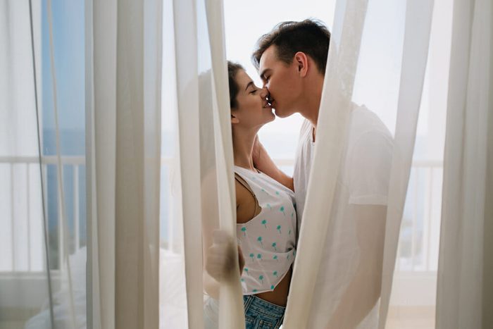 Graceful girl in white tank-top gently kissing her brunette boyfriend hiding behind light curtains. Portait of romantic young couple spending time together on balcony enjoying each other.