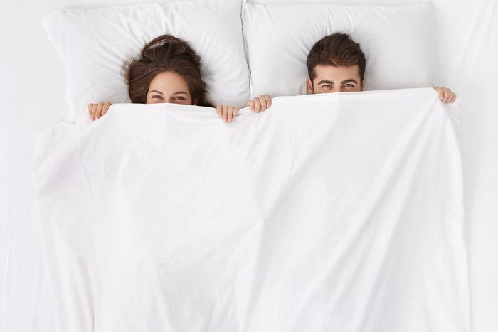 Funny married couple lying in bed and hiding under white blanket, looking at camera with eyes full of joy. Attractive Caucasian man and woman having fun in bedroom. Love and happiness concept