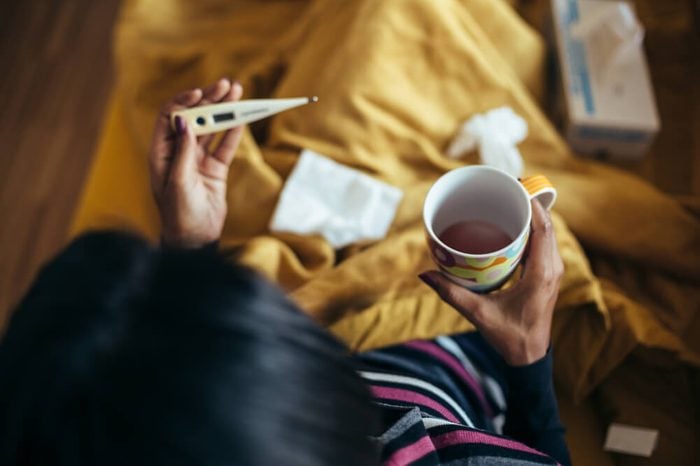 Sick woman drinking tea holding thermometer on couch