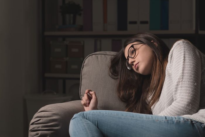 Sad young woman with glasses sitting on the couch at home, she is depressed and lonely