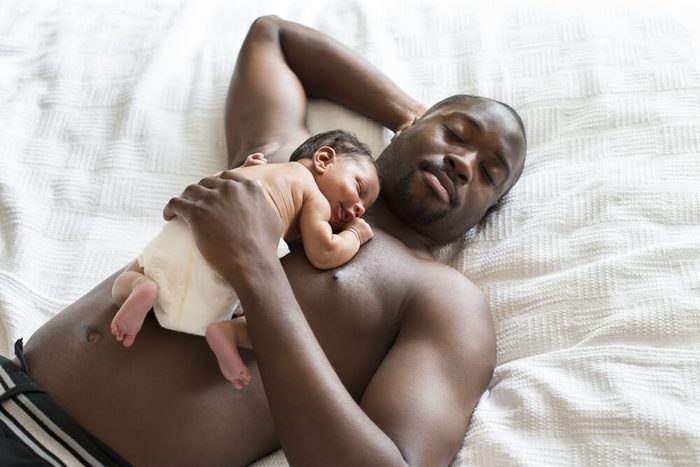A bare chested male lying down with his newborn sleeping baby on his chest.