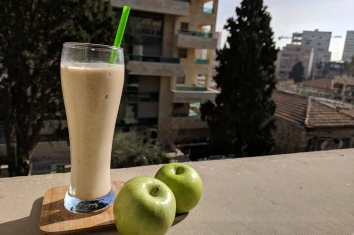 tall glass with smoothie next to two green apples on a ledge outdoors