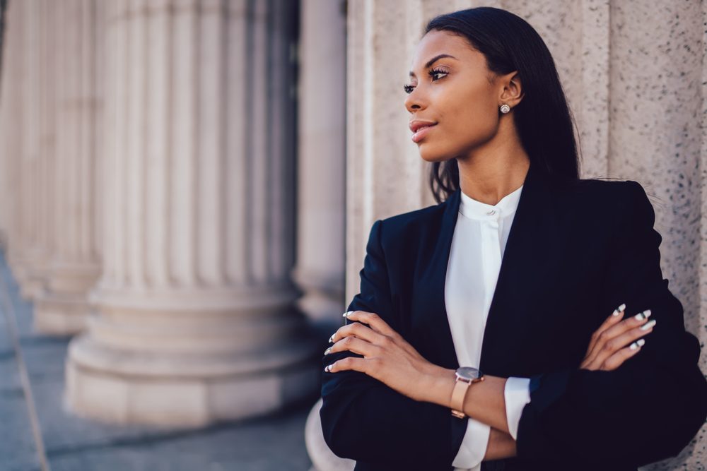 Female African American banker dressed in elegant black suit folding hands and looking on side standing against office building. Confident female entrepreneur with crossed arms looking on copy space