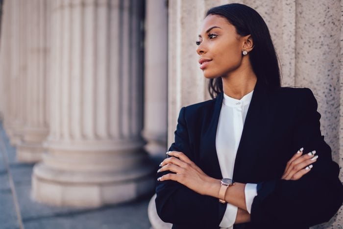 Female African American banker dressed in elegant black suit folding hands and looking on side standing against office building. Confident female entrepreneur with crossed arms looking on copy space