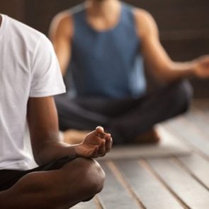 Young black man and a group of young sporty people practicing yoga lesson, sitting in Sukhasana exercise, Easy Seat pose, working out, indoor close up focus on mudra gesture, studio room