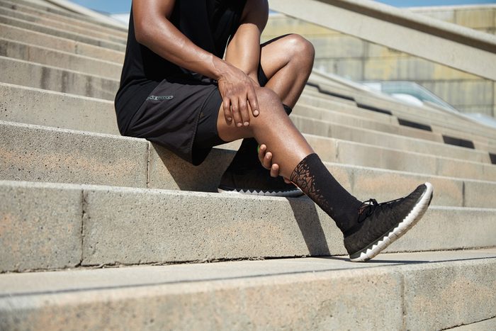 Sports injury concept. Cropped portrait of black male runner wearing black training outfit touching his leg in pain with clasped hands, having sprain or twitch in his knee after running exercises