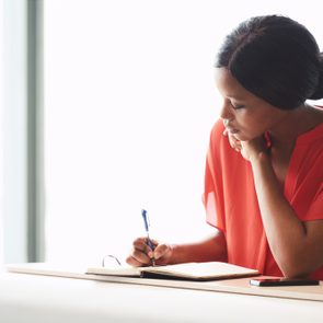 Self employed black female entrepreneur busy writing in her notebook while seated at a desk with a large bright window behind her in the background.
