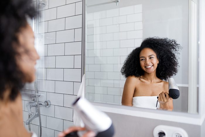 Smiling woman holding a hair dryer, looking at mirror on her blowing hair