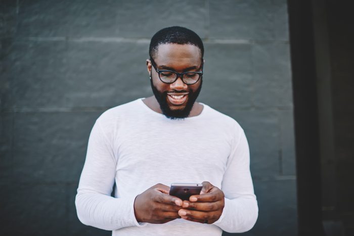 Cheerful african american man in spectacles searching cafes and restaurants in application on mobile,smiling dark-skinned guy reading news from networks on phone standing on copy space for advertising