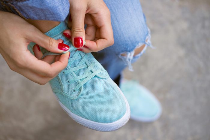 Female hands with a red manicure knotted laces on sports shoes. Young woman in blue jeans walking outdoors when she untied shoelace. A walk in the city.