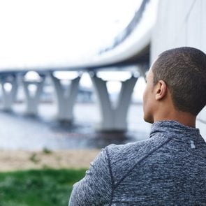 Rear view of thoughtful African American male runner in stylish sports jacket leaning on wall outdoors, looking into distance at sea, relaxing mind or meditating after running exercise in the morning